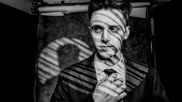 Man in business suit with dramatic window-blind shadows cast across his face and torso.