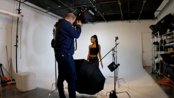 Photographer operating camera on tripod while model in black outfit poses during studio fashion shoot.