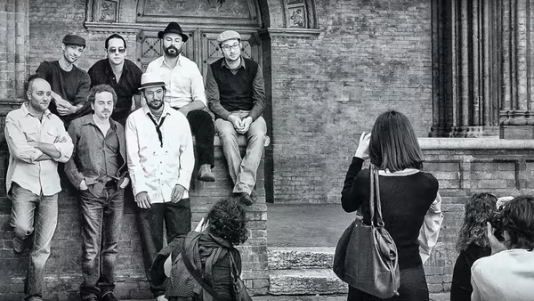 Group of men leaning against brick wall while being photographed by woman in foreground.