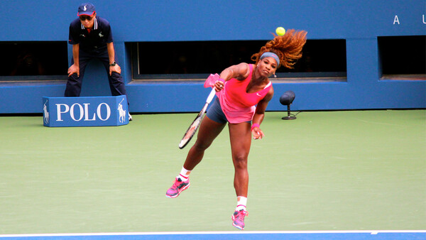 Tennis player in pink outfit executing a serve on a hard court with an umpire seated in the background.