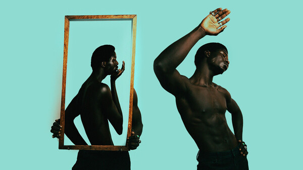 Shirtless man holding wooden frame mirror showing reflection against turquoise background.