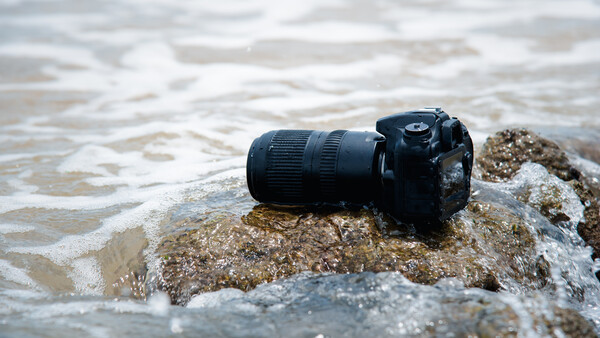 Black DSLR camera with telephoto lens resting on a rocky outcrop in shallow ocean waves.