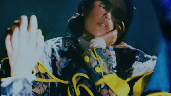 Person in yellow and black patterned jacket against blue background, hand raised expressively.