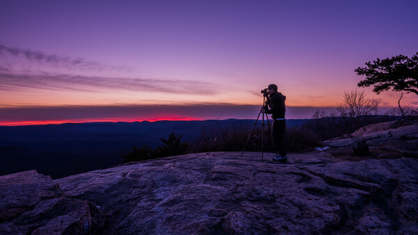 Photographer on rocky outcrop silhouetted against vibrant purple and pink sunset sky.