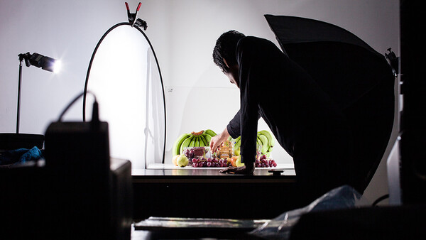 Photographer setting up a still life shot of fresh fruits under studio lighting with reflectors and key light positioned around the setup.