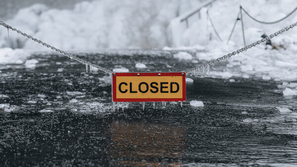 Red and yellow CLOSED sign suspended by chains over an icy river with snow-covered banks.