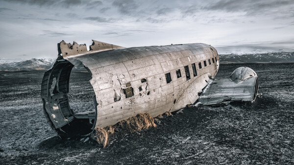 Abandoned aircraft fuselage lies on dark volcanic sand with mountains in the distance under overcast sky.