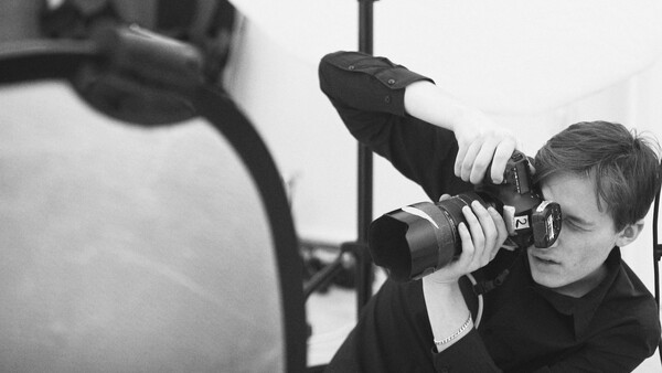 Photographer holding a telephoto lens with sunglasses, composing a shot through the viewfinder in black and white.