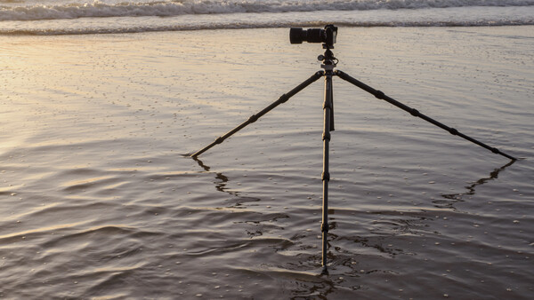 Tripod with inverted legs planted in shallow water at a beach during golden hour.
