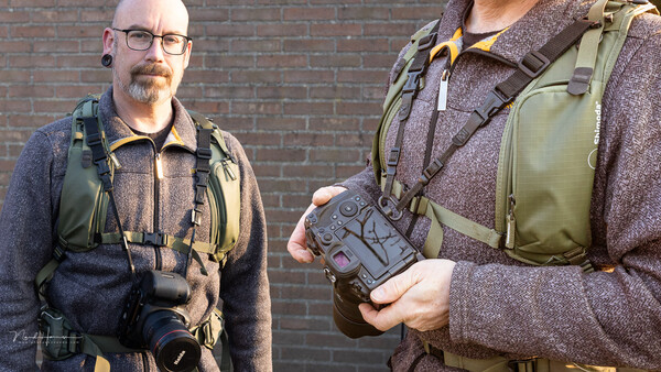 Two photographers wearing tactical vests and backpacks stand against a brick wall, one holding a camera and the other holding a gimbal stabilizer.