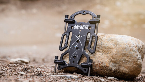 Black phone mount holder with grip arms positioned against a large stone on sandy ground.