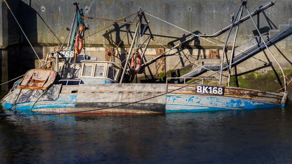 Weathered fishing vessel with registration number BK168 moored against a concrete seawall, its tangled rigging and nets silhouetted against gray water.