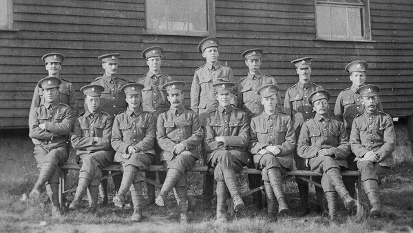 Fifteen uniformed soldiers arranged in three rows posing before a wooden building.
