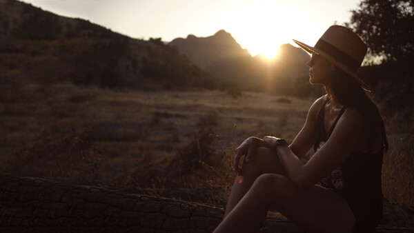 Woman wearing a hat sitting outdoors during golden hour sunset, gazing toward distant mountains.