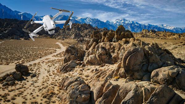 White drone hovering over rocky desert terrain with snow-capped mountains in the background.