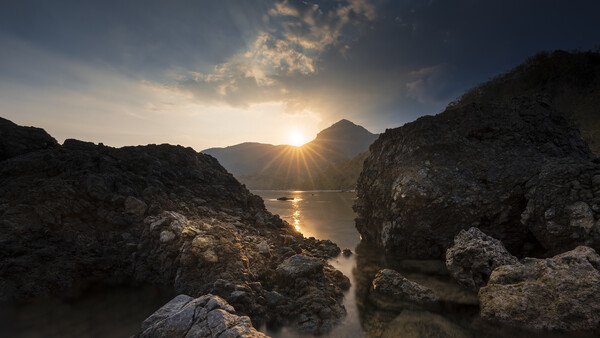 Rocky coastline framing a mountain and sun at golden hour with calm water between dark stone formations.