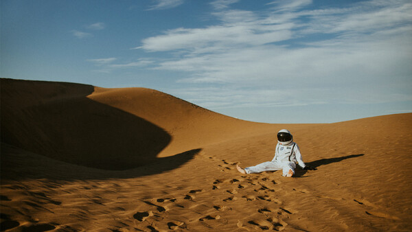 Astronaut in white spacesuit sitting alone on golden sand dune with footprints visible behind.