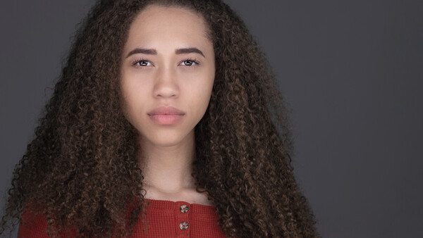 Portrait of a young woman with long curly dark hair wearing a red ribbed top against a neutral gray background.
