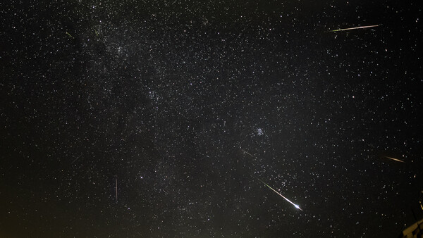 Milky Way galaxy stretching across night sky with multiple meteor streaks and silhouetted mountain ridge below.