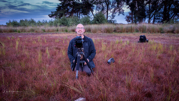 Photographer holding a camera on tripod in a field of reddish grasses at dusk.
