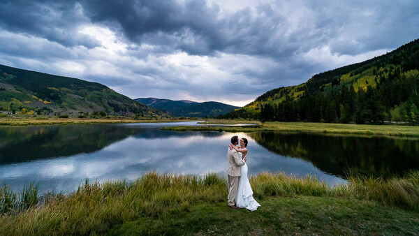 Couple in wedding attire standing in grass by a calm lake surrounded by forested mountains under dramatic cloudy sky.