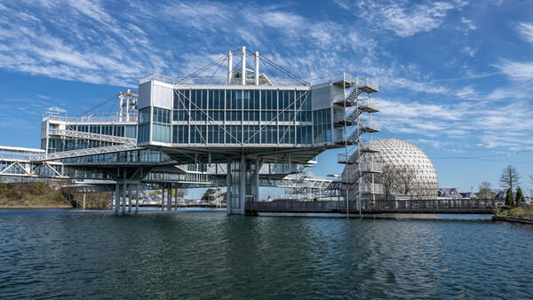 Modern waterfront building with white vertical columns and glass facade cantilevers over calm blue water.