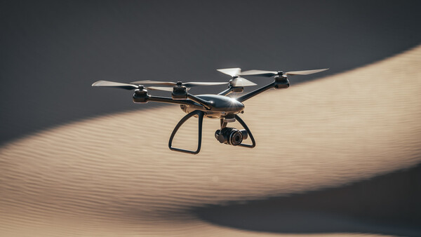 Quadcopter drone hovering above rippled sand dunes with dramatic side lighting casting sharp shadows.
