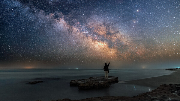 Silhouetted figure standing on a rocky outcrop facing a vast starry night sky with the luminous Milky Way galaxy arching overhead.