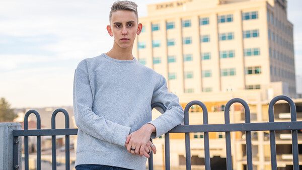 Young man in light blue sweatshirt leaning against railing with multi-story building behind him.