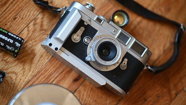 Vintage rangefinder camera with chrome and black body on wooden surface next to film and lens.