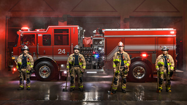 Four firefighters in yellow-and-black turnout gear stand in front of red fire trucks in a station bay with dramatic red lighting.