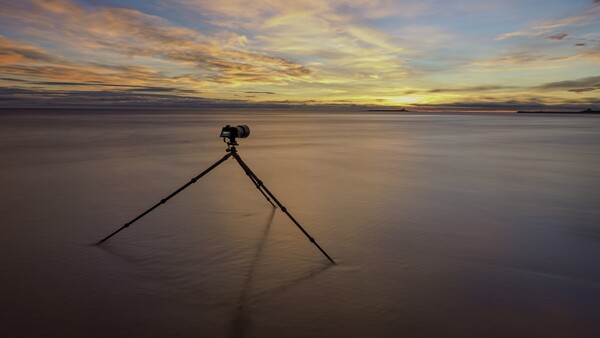 Tripod standing alone in shallow water at sunset with golden sky reflected below.