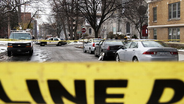 Yellow crime scene tape in sharp focus with urban street scene and parked vehicles blurred in background.
