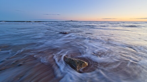 Rocky outcrop surrounded by flowing seawater during twilight, with soft waves creating circular patterns around the stone.
