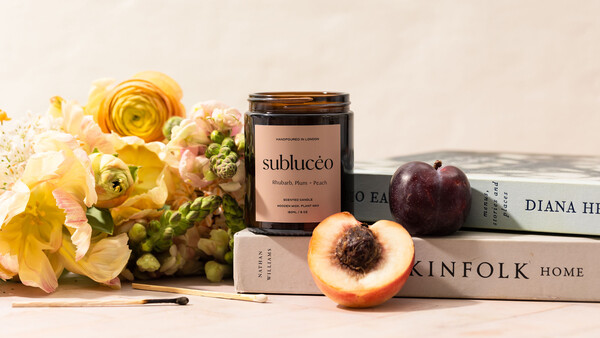 Styled flat lay with yellow flowers, cosmetic jar, sliced peach, dark plum, and stacked books on neutral background.