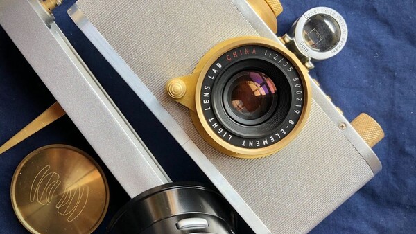 Vintage rangefinder camera with white leather strap and brass fittings displayed with accessories on blue fabric.