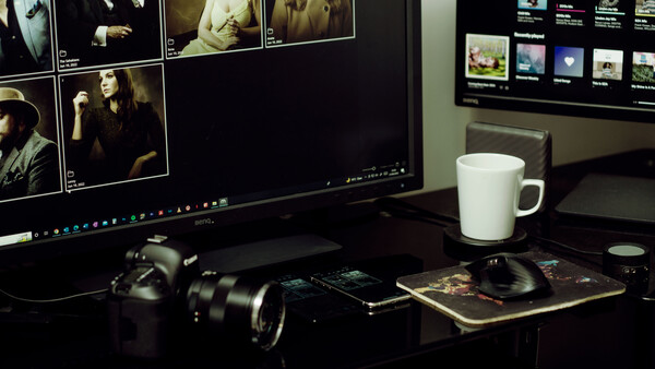 Photographer's desk setup with DSLR camera, lens, and monitor displaying photo editing software.