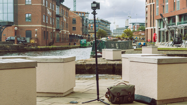 Camera on tripod positioned at waterfront location with brick buildings and modern development in background.