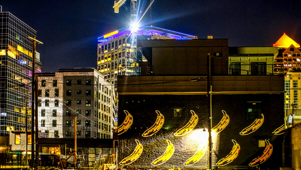 Nighttime urban street with illuminated banana murals on dark building facade surrounded by lit skyscrapers.