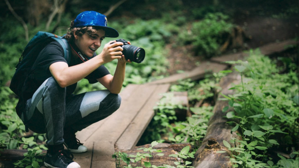 Photographer crouching on a wooden boardwalk through a forest, holding a camera to their eye while composing a shot.