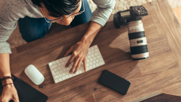 Photographer working at a wooden desk with camera equipment and computer peripherals.