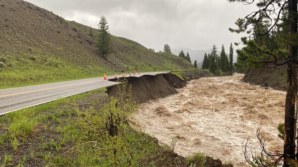 Swollen river with muddy water rushing past a damaged highway embankment during flood conditions.