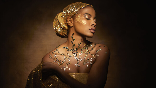 Woman with gold leaf makeup and glittering body art against warm brown background.