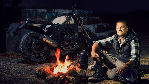 Man crouching by a campfire next to a motorcycle at night.