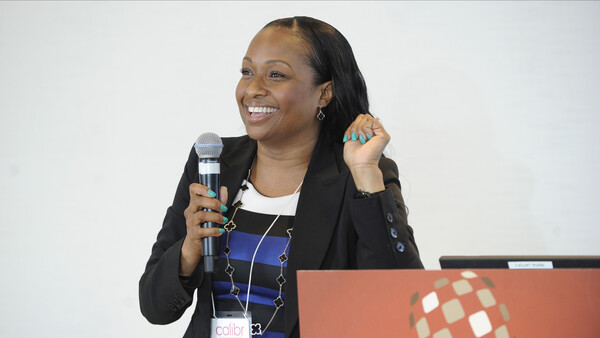 Woman speaking at a conference with microphone, wearing black blazer over striped top.