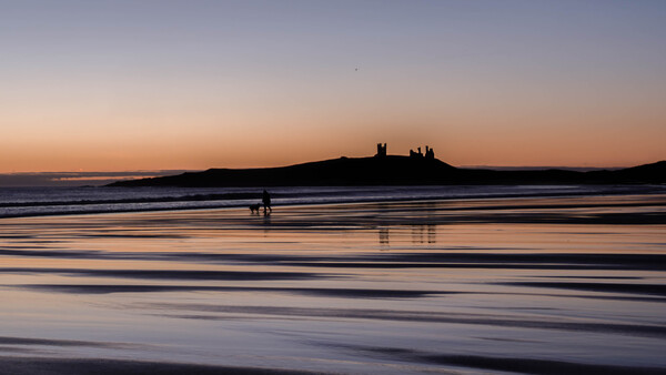 Silhouetted castle ruins on a distant headland at sunset, with a lone surfer in the shallow foreground water.