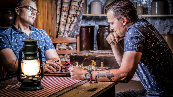 Two men playing chess by lamplight in a rustic interior setting.