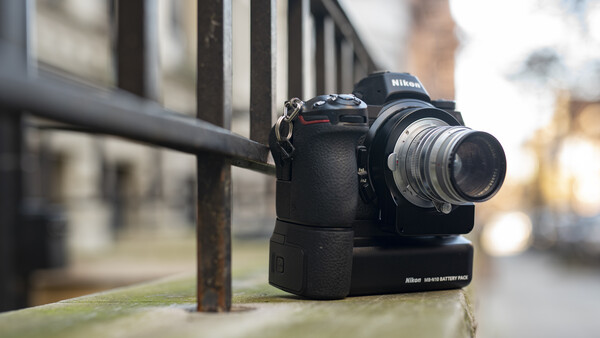 Nikon DSLR camera with telephoto lens mounted on a battery grip, resting on a ledge against an urban backdrop.