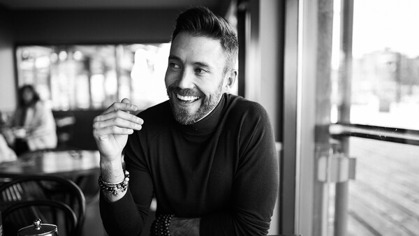 Man with beard smiling at camera in bright indoor space with windows, black and white portrait.