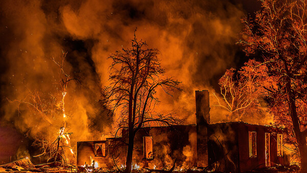Residential structure engulfed in intense flames at night, surrounded by bare trees silhouetted against orange and black smoke.